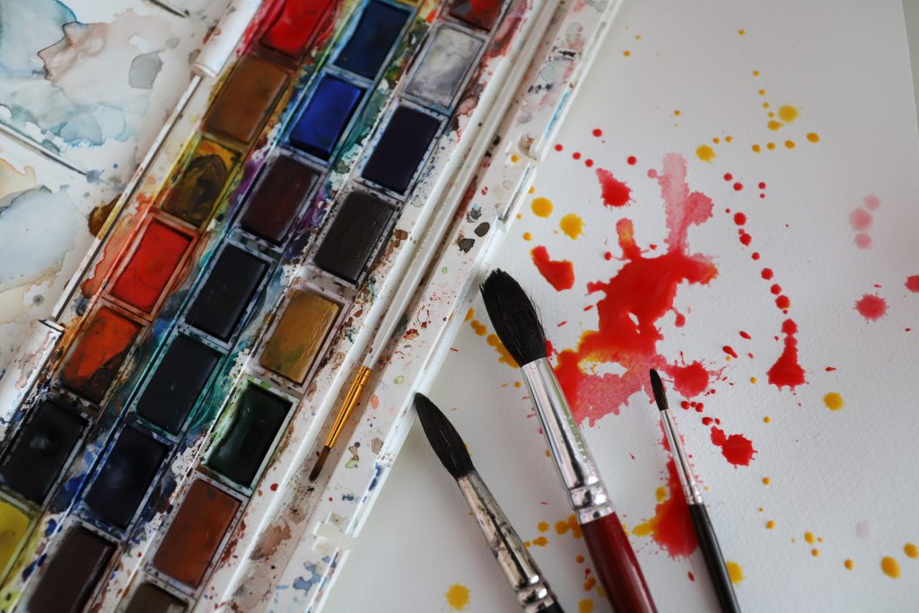 Kuvituskuva. Akvarellivärit, pensselit ja vesiväriroiskeita